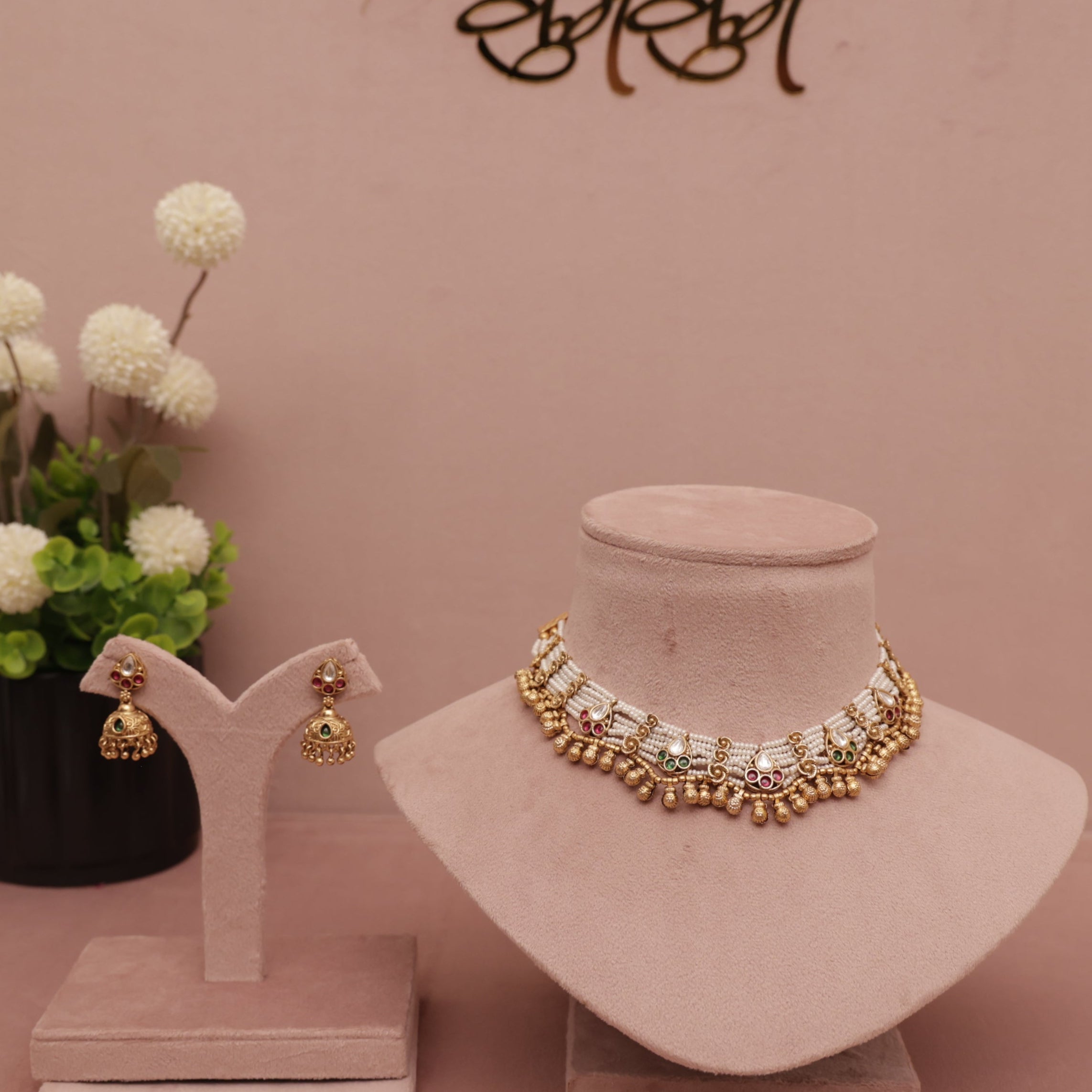Gold necklace and earrings on display with a vase of flowers and 'BIBI' branding in the background.