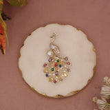 Decorative peacock brooch with gemstones on a round white plate with gold rim against a pink background.