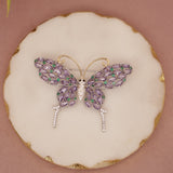 Butterfly-shaped jewelry piece with purple and green gemstones on a beige background
