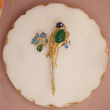 Gold bird-shaped brooch with green and blue stones on a white cushion with gold border