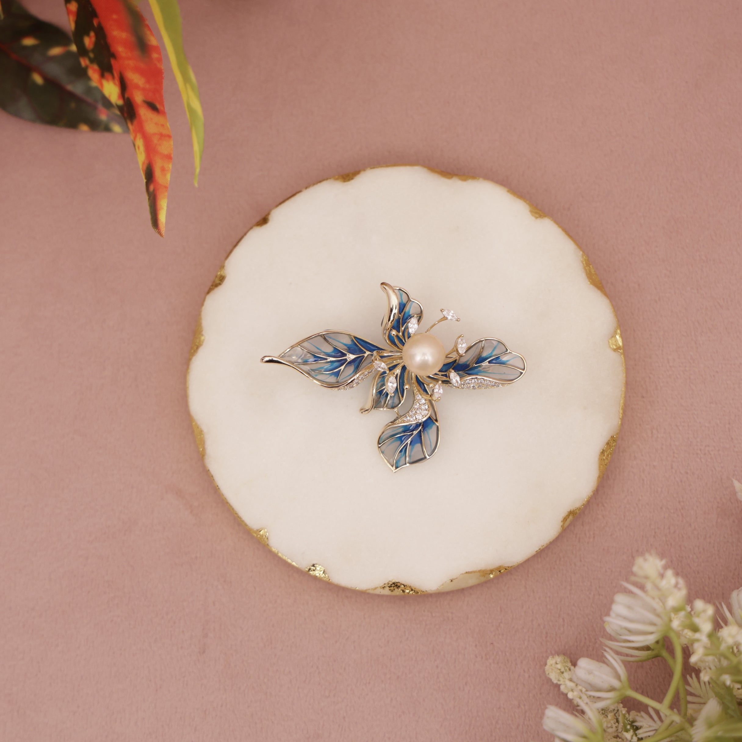 Decorative brooch with pearl and blue leaves on a white round base against a pink background