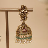Gold-toned traditional Indian jhumka earrings with a deity figure at the top, green bead accents, and small pearl-like dangling beads, displayed on a stand against a warm beige background.
