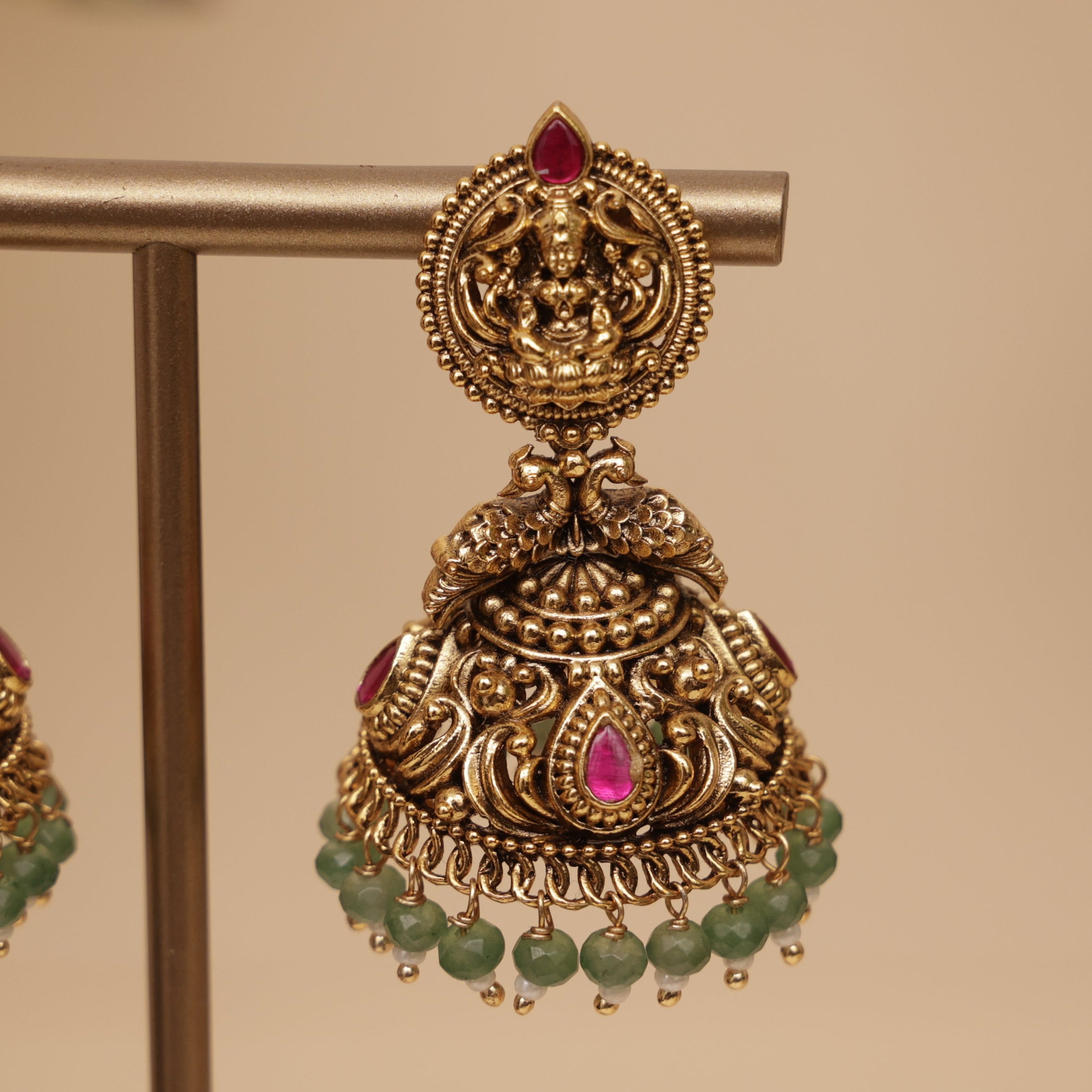 An ornate gold-toned traditional Indian jhumka earring displayed on a metal stand, featuring a detailed filigree top with a small red stone, a teardrop-shaped pink stone, and dangling green bead chains.