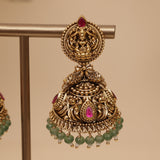 An ornate gold-toned traditional Indian jhumka earring displayed on a metal stand, featuring a detailed filigree top with a small red stone, a teardrop-shaped pink stone, and dangling green bead chains.