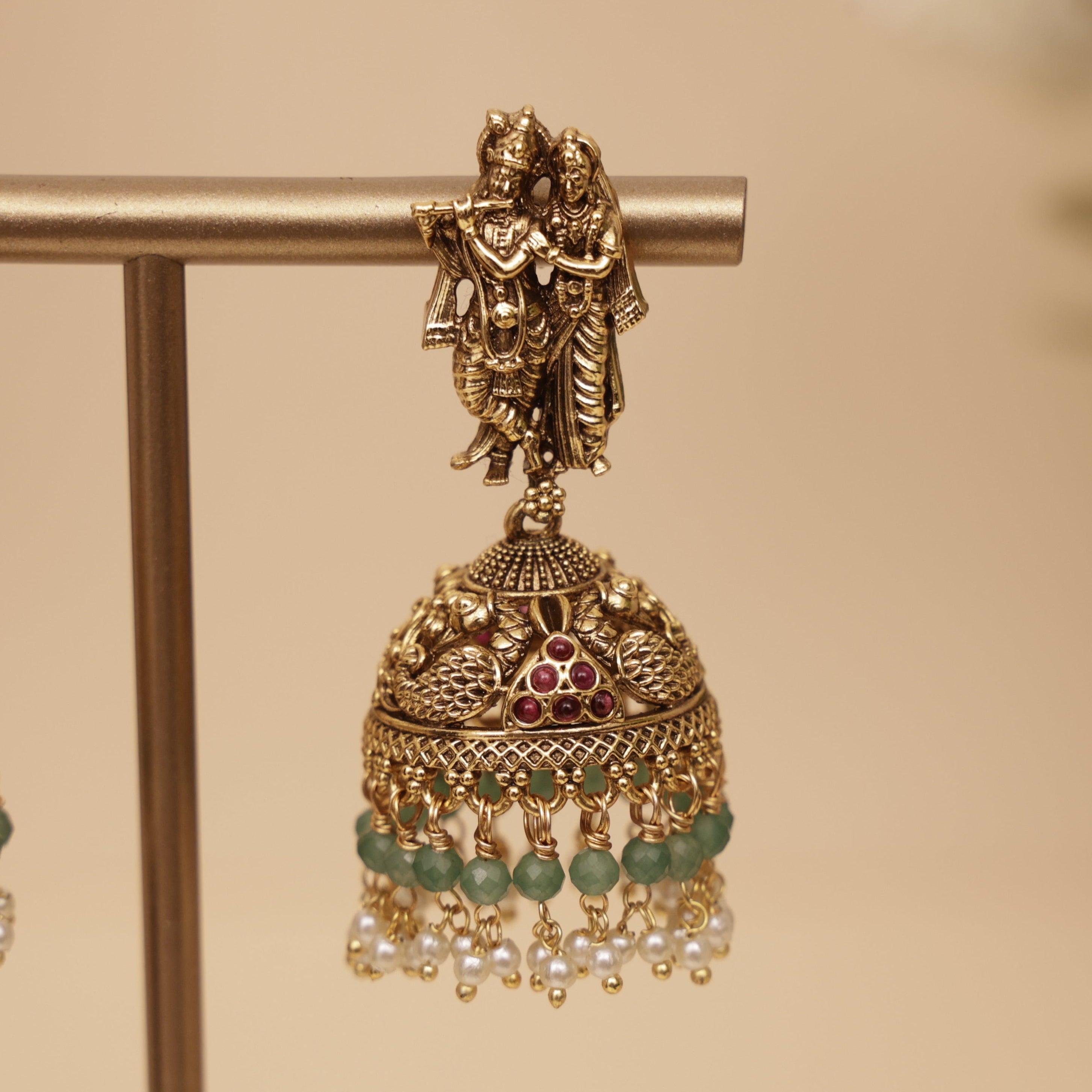 Gold-toned traditional Indian jhumka earrings with a deity figure at the top, green bead accents, and small pearl-like dangling beads, displayed on a stand against a warm beige background.